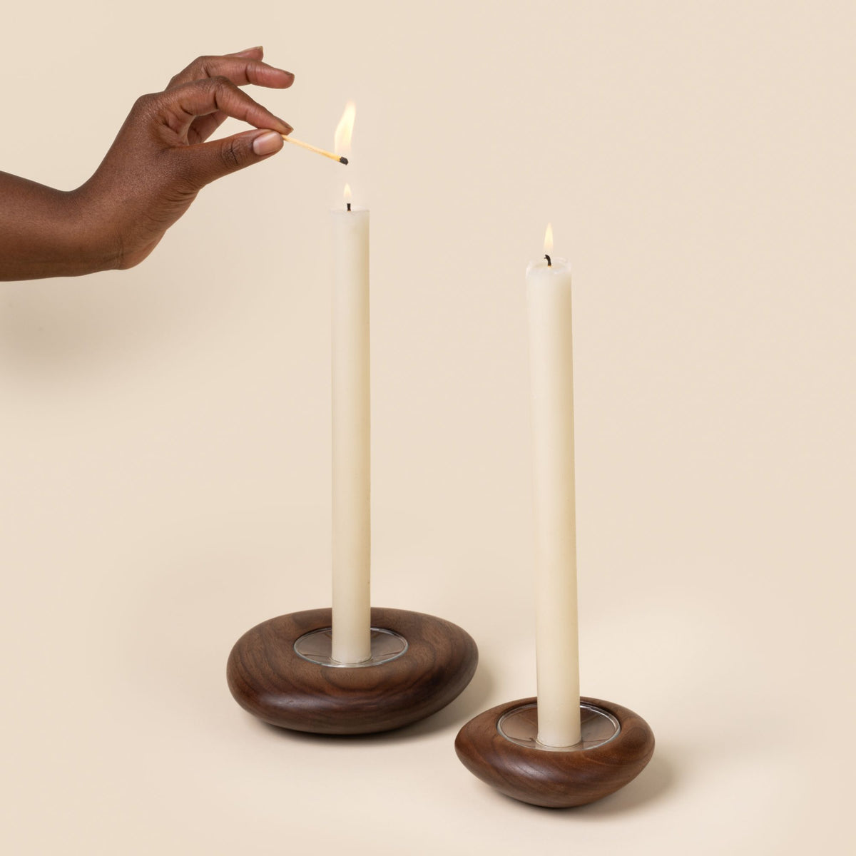 A hand lights two lit white taper candles in round, light wood candle holders with visible grain, displayed against a neutral background.