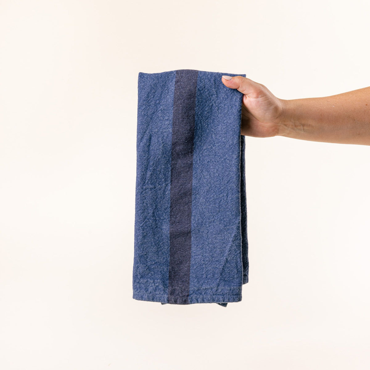 A hand holds a folded linen towel in an indigo color with a plum stripe in the middle