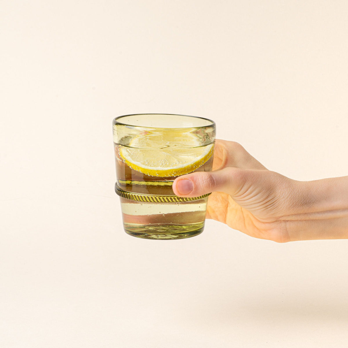 A hand holding an olive short recycled glass tumbler with a ridged band detail, filled with water and a lemon slice.