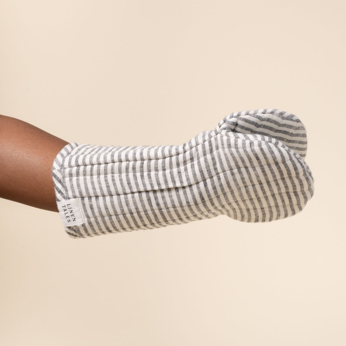 Hand inside a gray-and-white striped linen oven mitt with stitched channels and a small ‘Linen Tales’ label, against a light background.