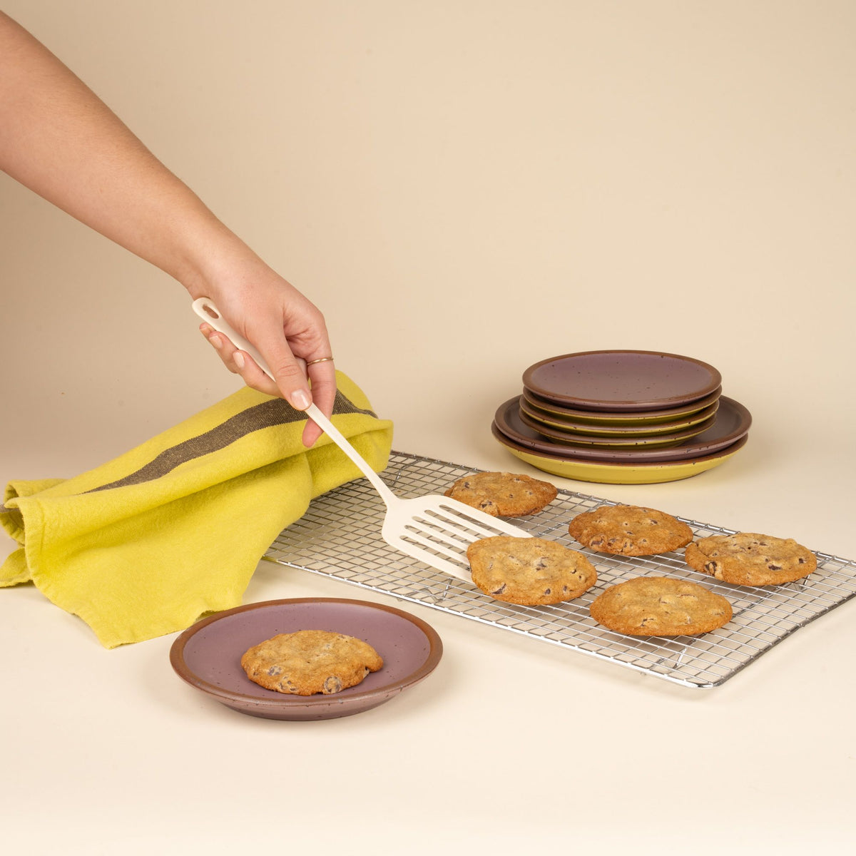 A hand uses a white enamel spatula to lift cookies off a baking tray, surrounded by soft purple plates, and a bright yellow towel.