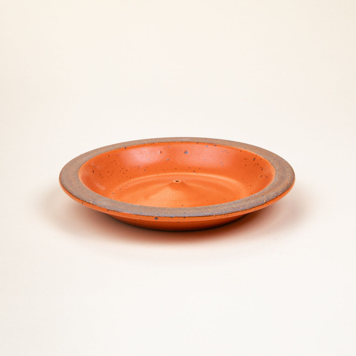 A ceramic plate-shaped incense stick holder with an unglazed rim and glazed in a bold orange color featuring lots of iron speckles.