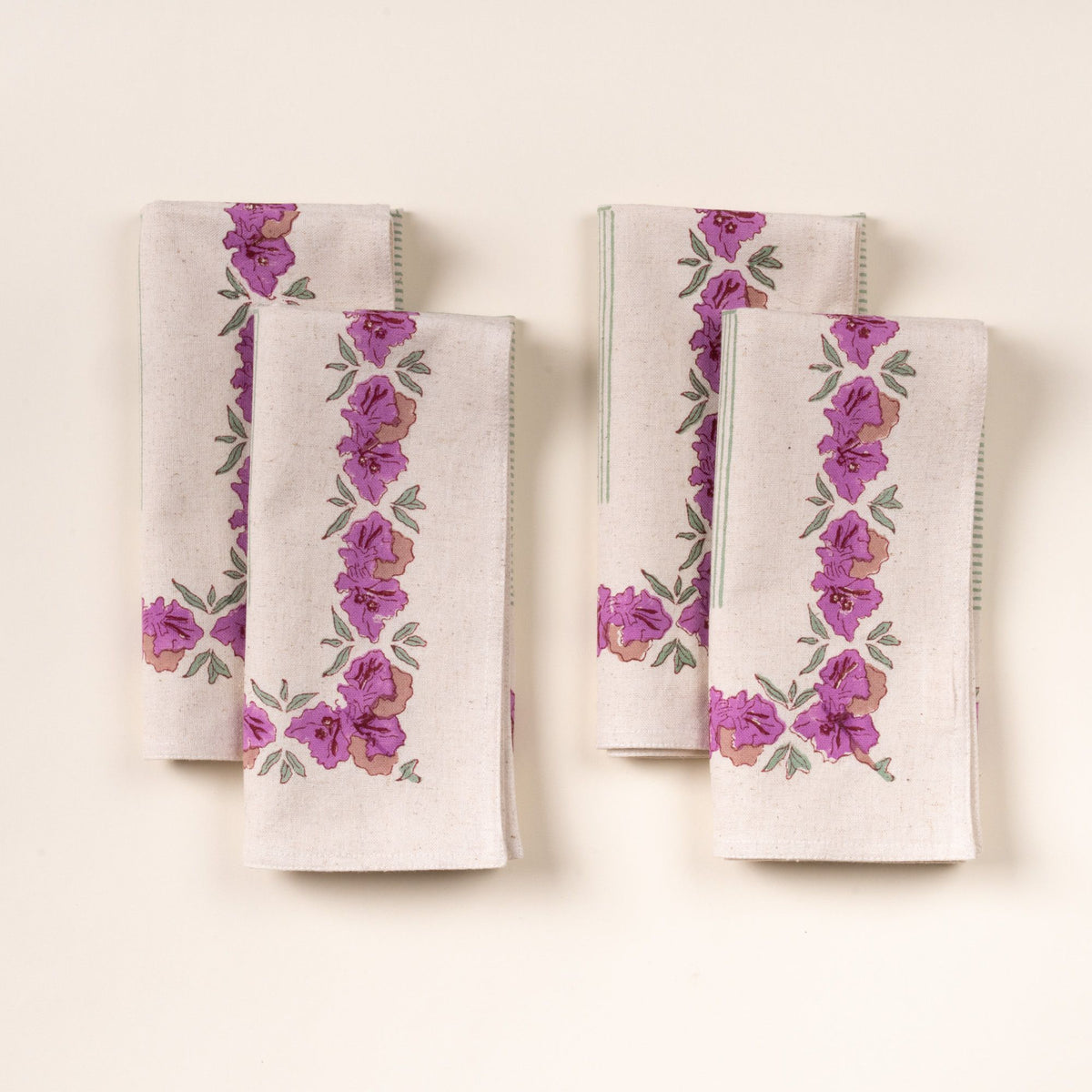 Four folded dinner napkins with purple floral borders and green stitched edges, arranged in two pairs on a cream background.
