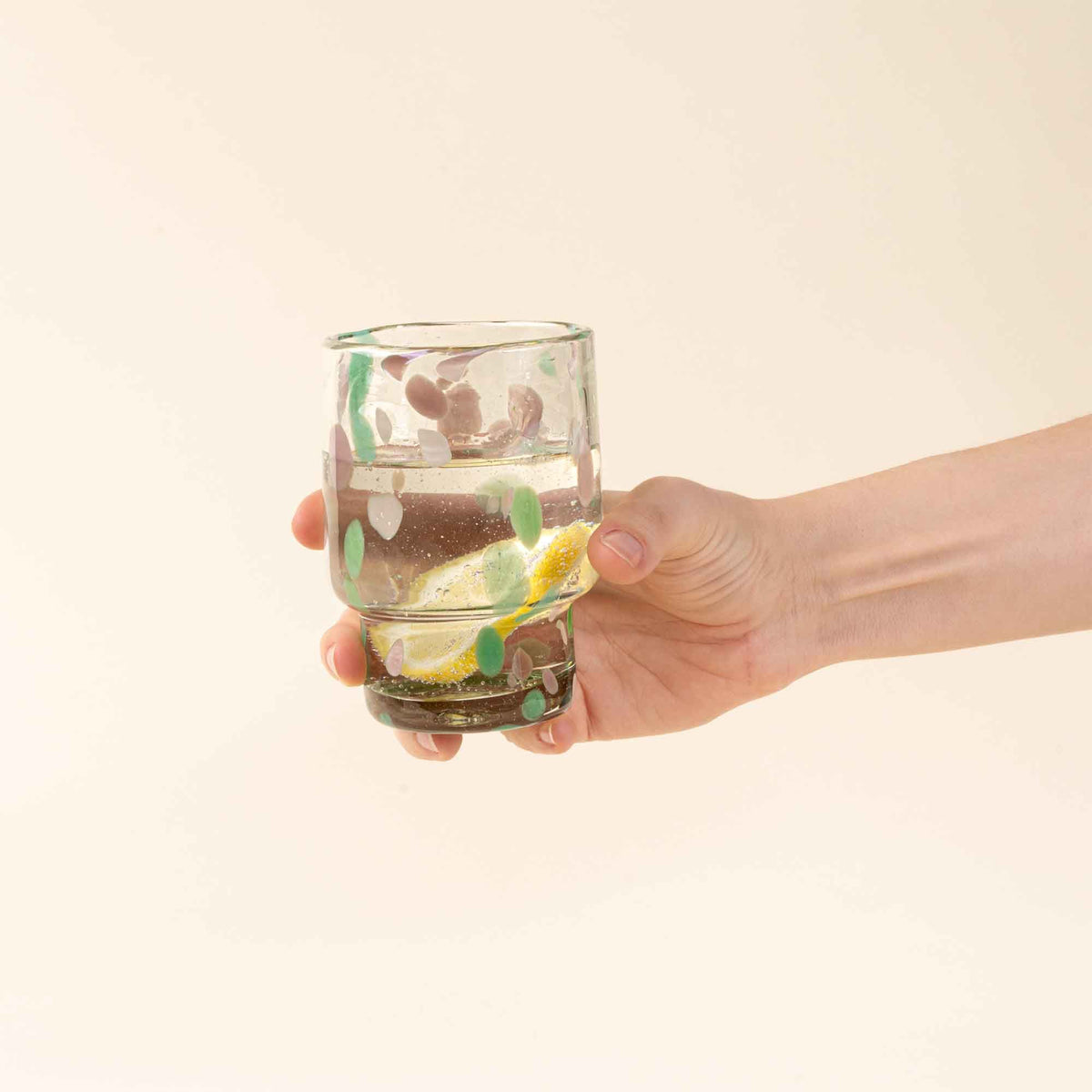 A hand holds a short clear glass with pastel green and pink speckled patterns, filled with water and lemon slices.