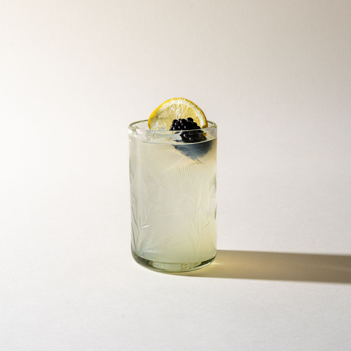 A clear tumbler glass made of recycled glass with etched thistle designs on it, filled with a cocktail and topped with a lemon slice and blackberry.