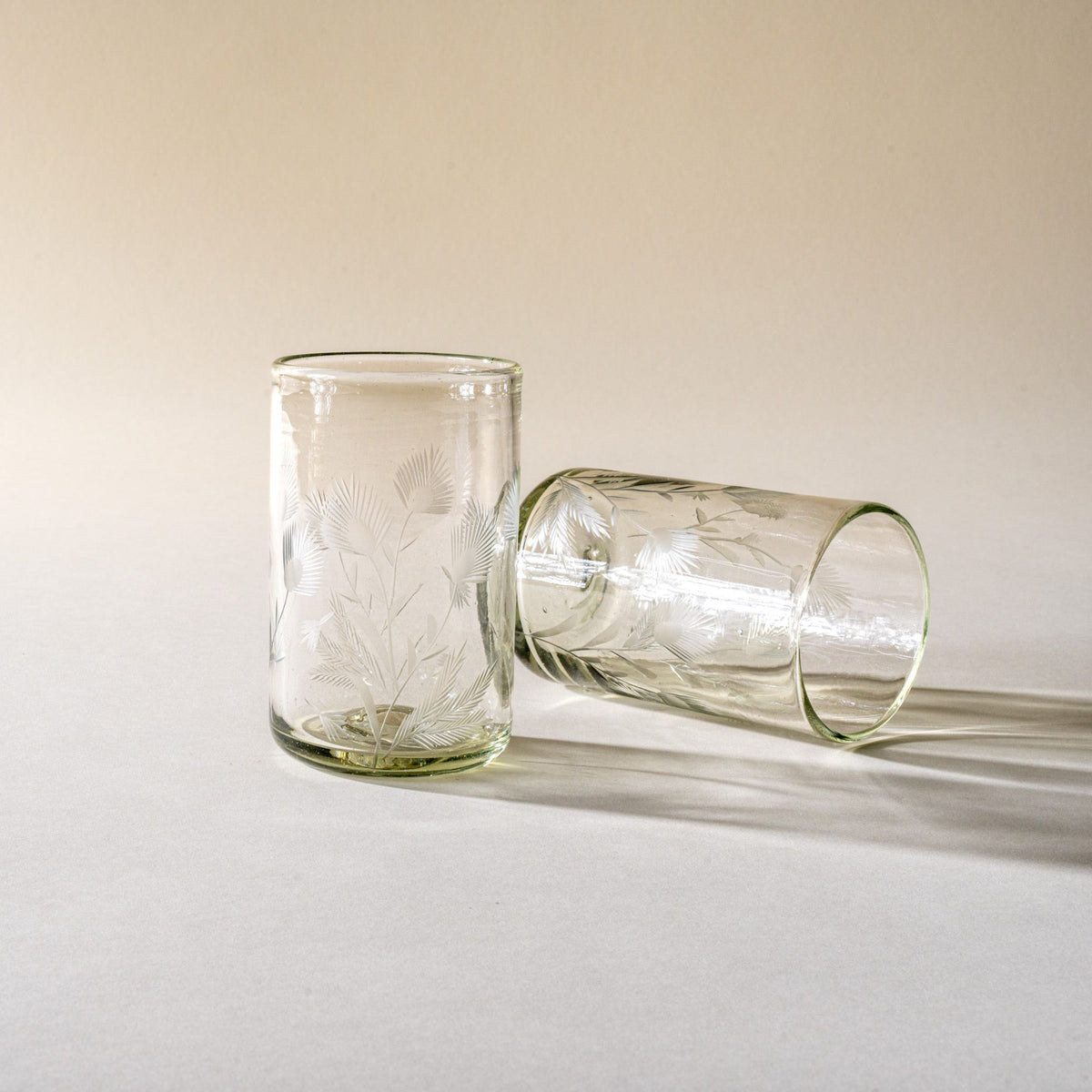 Two clear tumbler glasses made of recycled glass with etched thistle designs on it, one standing up, one laying on the side.