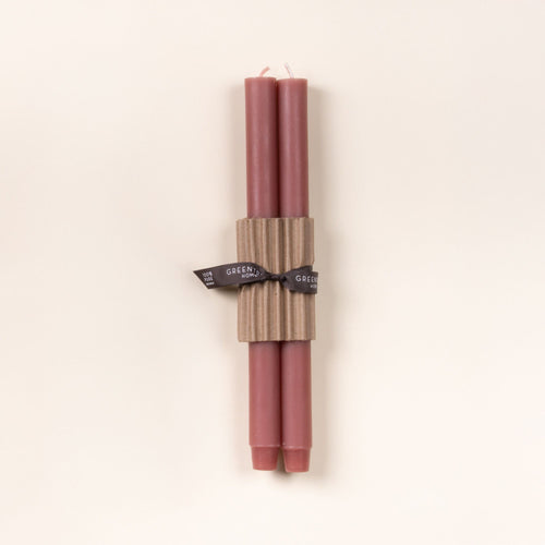 Two dusty mauve pink taper candles bundled with corrugated cardboard and tied with a black ribbon on a neutral background.