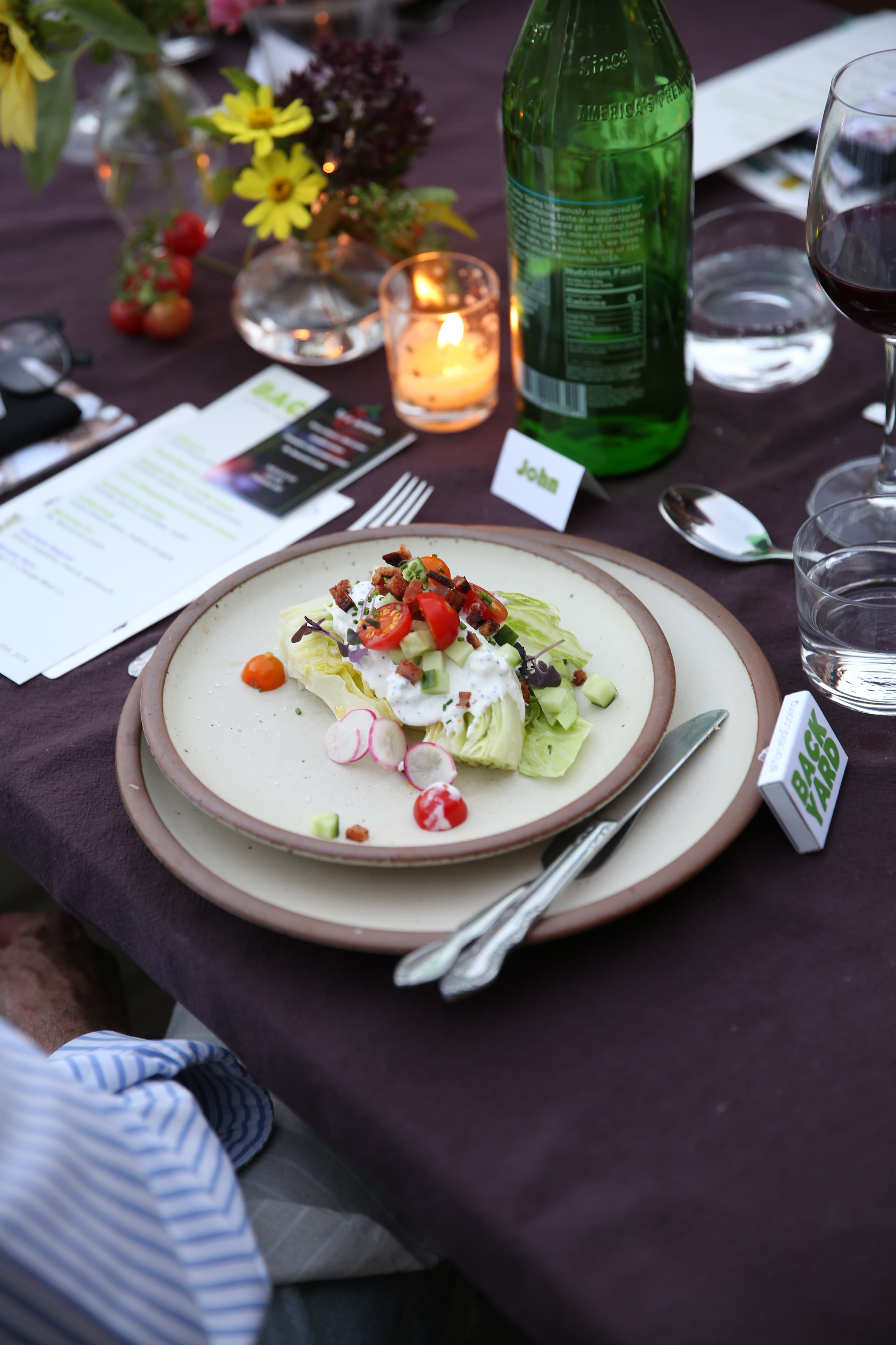 A place setting at an outdoor table with warm off-white ceramic plates, silverware, candles, and drinking glasses.
