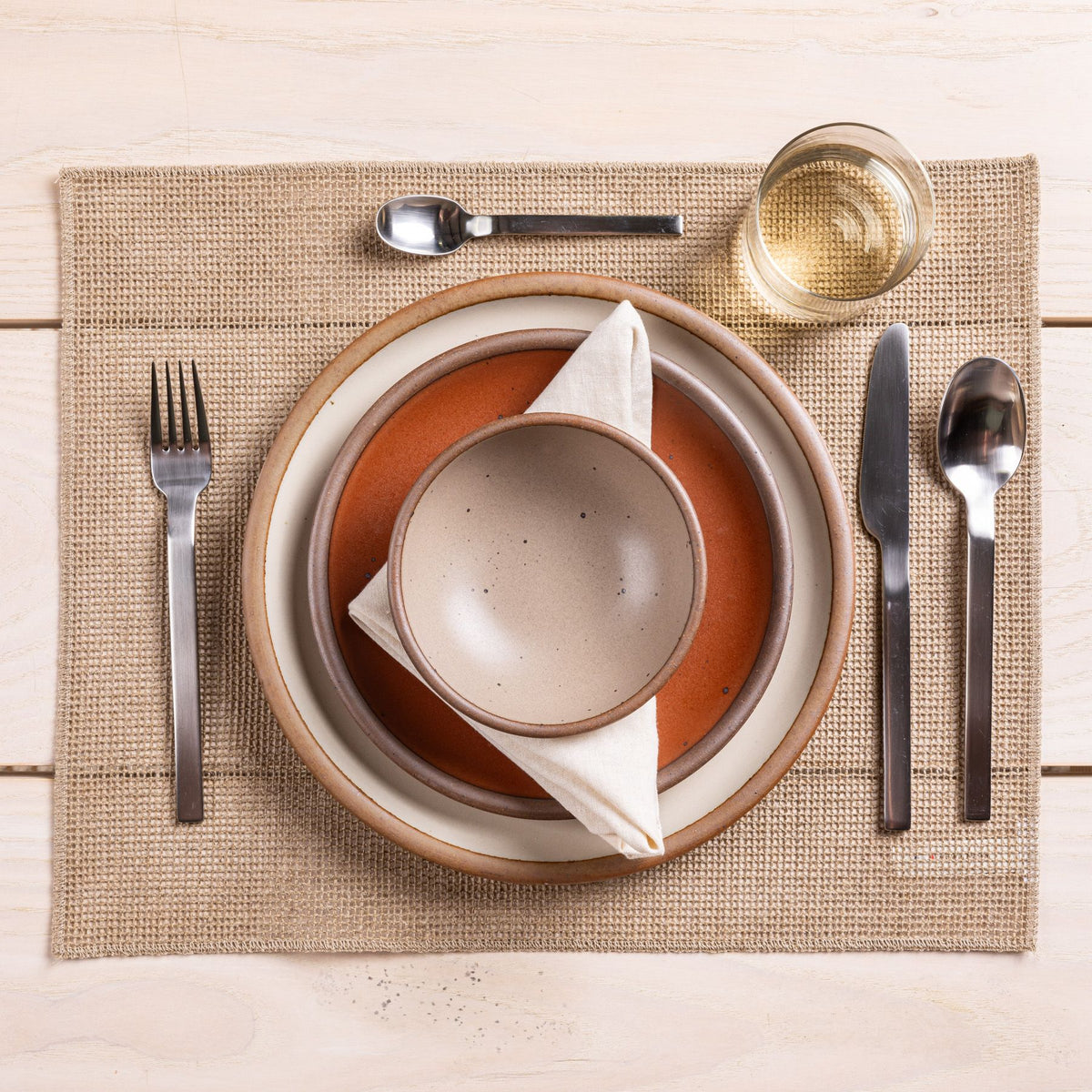 A table setting featuring ceramic plates, a woven placemat, a glass, and steel flatware