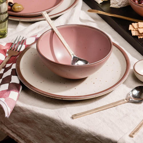 Pink bowl with a spoon resting inside, set on a speckled plate on a linen tablecloth with brass and steel flatware utensils and napkins nearby.