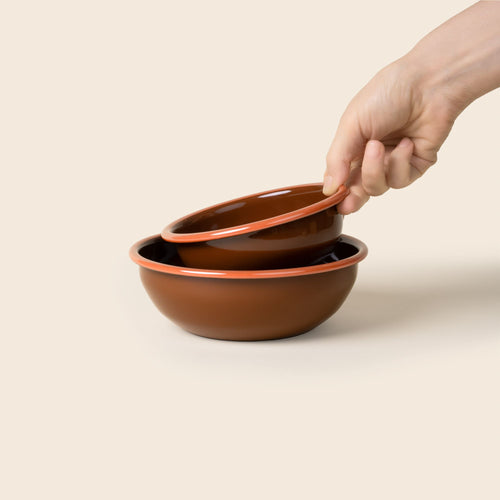 A hand holds a small enamel bowl to nest in a medium enamel bowl; both bowls are dark red-brown with an orange rim.