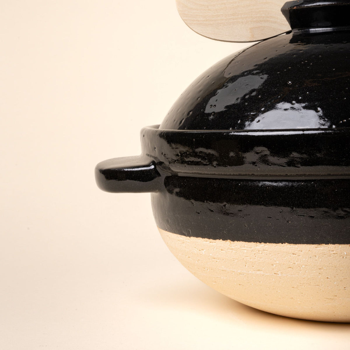 Closeup of the side of a black ceramic donabe pot with lid and a small wooden spoon on a light background.