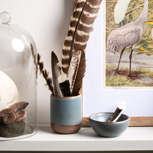 A small short ceramic cup in a blue-grey color with unglazed base and rim filled with bird feathers, on a shelf with a small tiny bowl.