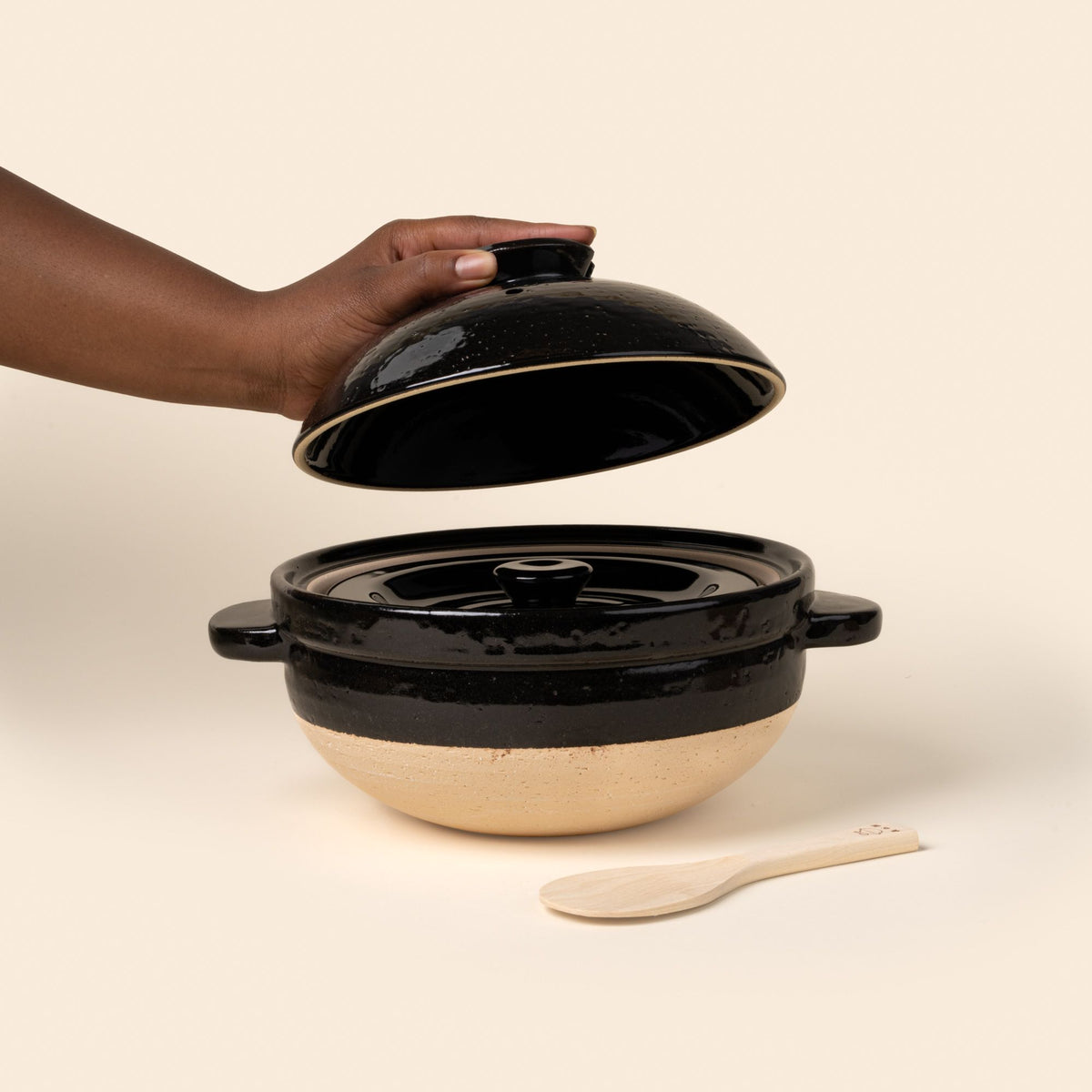 A black-and-neutral color claypot with handles and a shallow-dome shaped lid. The lid is being lifted off by a hand. The cooking vessel sits atop a wooden board next to a wooden spoon.