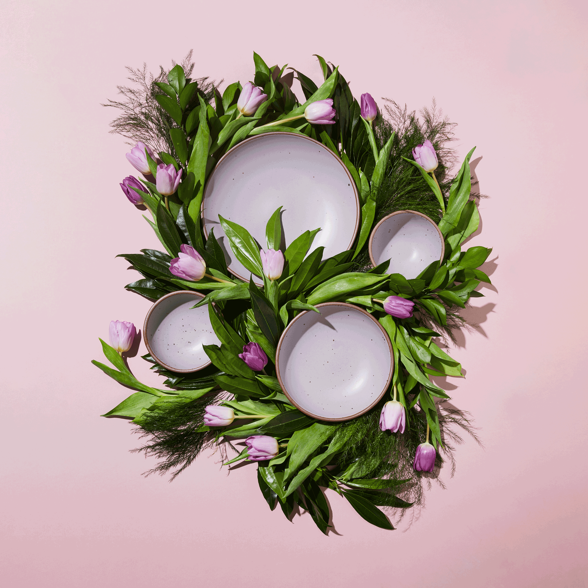 An overhead view of 4 plates and bowls of various sizes in pastel purple colors surrounded by purple tulips and greenery