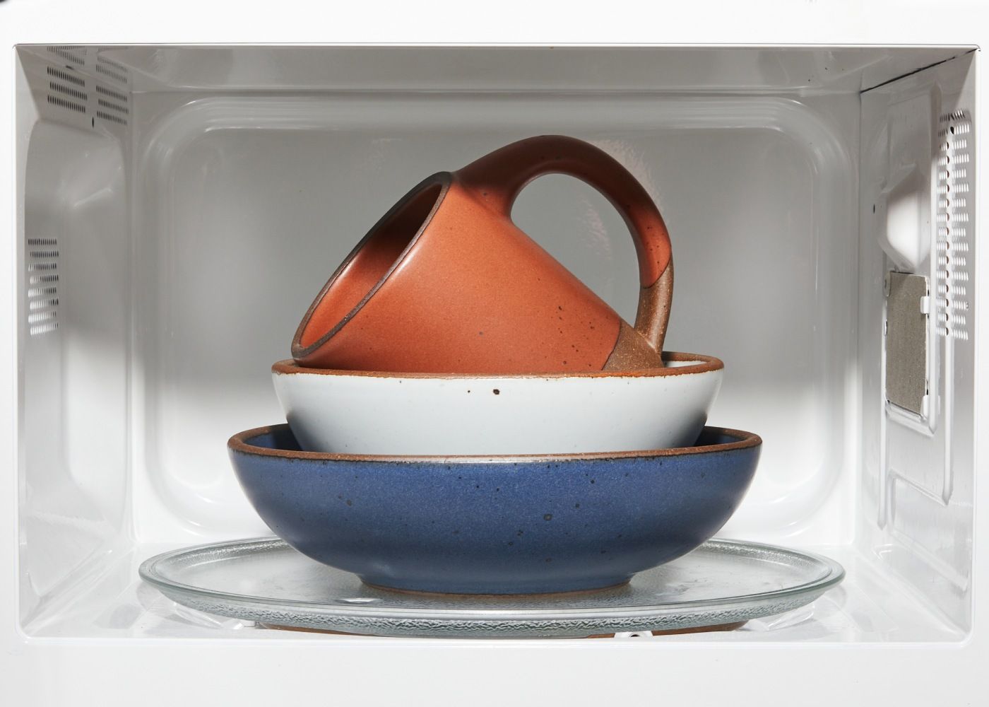 In a microwave sits a stack of a blue shallow bowl, a white ice cream bowl, and a cool terracotta mug