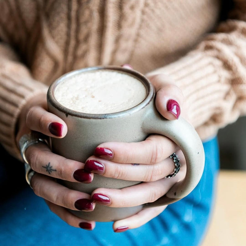Hands with burgundy nail polish hold a large ceramic mug filled with a frothy beverage, against a backdrop of a textured brown sweater and blue jeans.