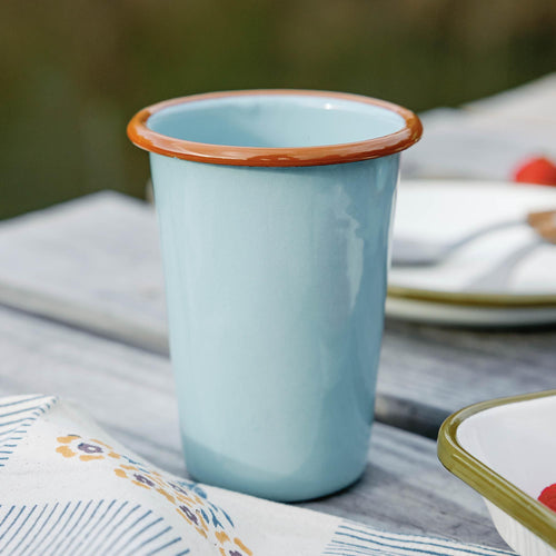 Light blue enamel tumbler with a brown rim on a wooden table, patterned cloth and dishes nearby.