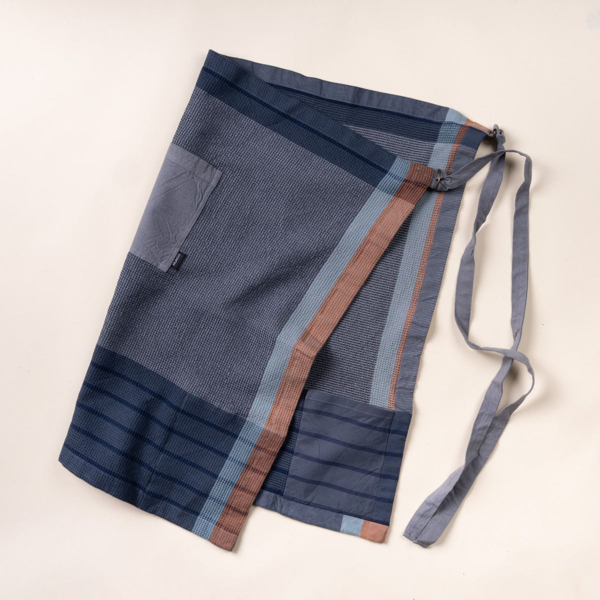 A folded woven kitchen apron in navy and slate blue with light blue and rust stripe accents, shown on a light background with a front pocket and long tie straps.