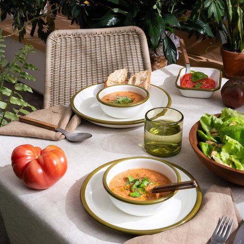 A table with enamel plates and bowls in a white color with olive green rim, green glasses, and more.