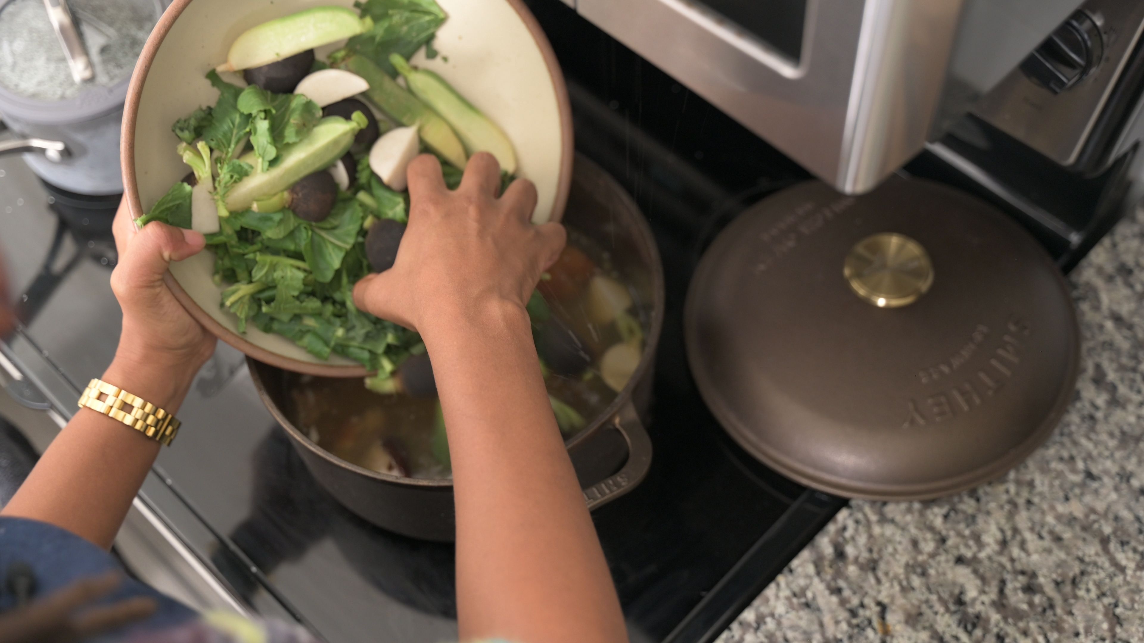 A hand holding a large bowl of vegetables with the other hand pouring them into a dutch oven.