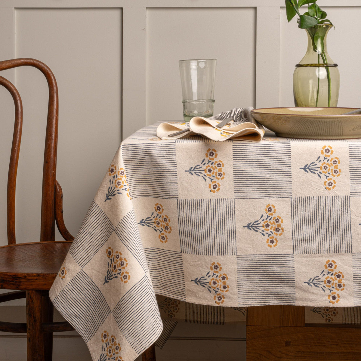 A blue-and-white checkered cloth tablecloth printed with small yellow flower bouquets on a table with grey-green pottery with green vase.