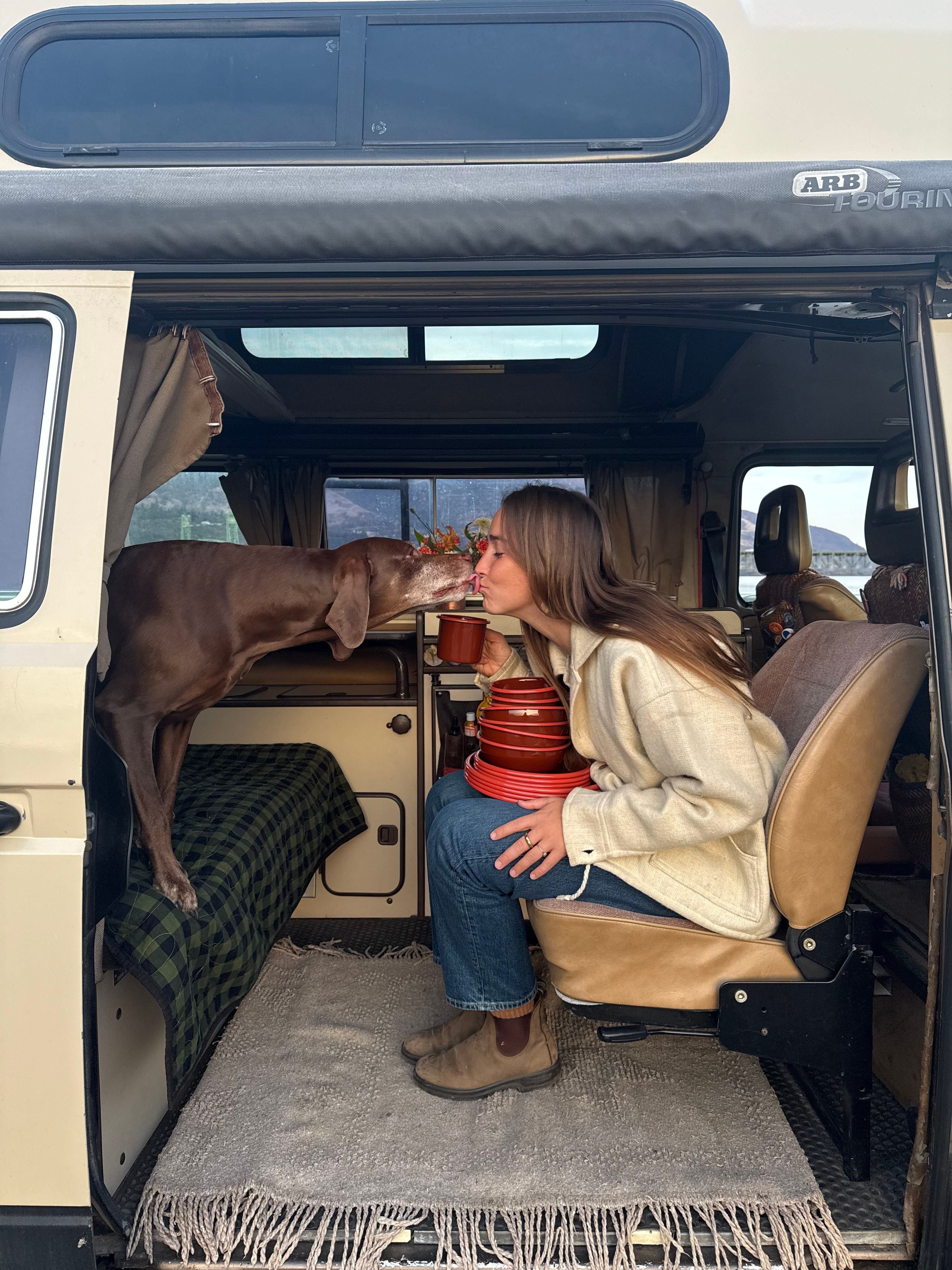 A girl and a dog smooch in a VW Vanagon. East Fork Enamelware sits on the girls lap.
