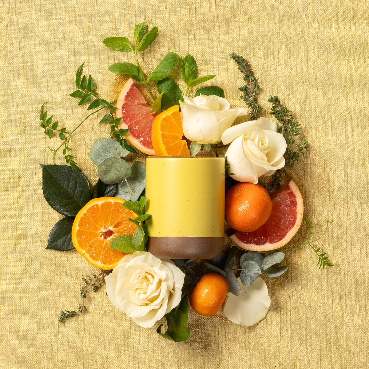 A large ceramic vessels in a zesty yellow color laying on its side in an artfully styled bed of roses, fruits, and greenery