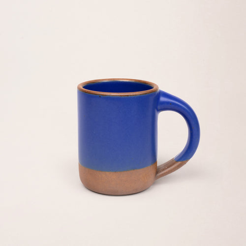 A medium sized ceramic mug with handle in a cobalt blue color featuring iron speckles and unglazed rim and bottom base.