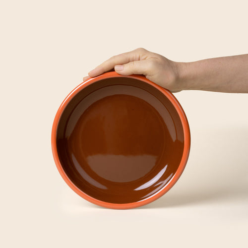 A hand props up an enamel side plate in brown-red with an orange rim.