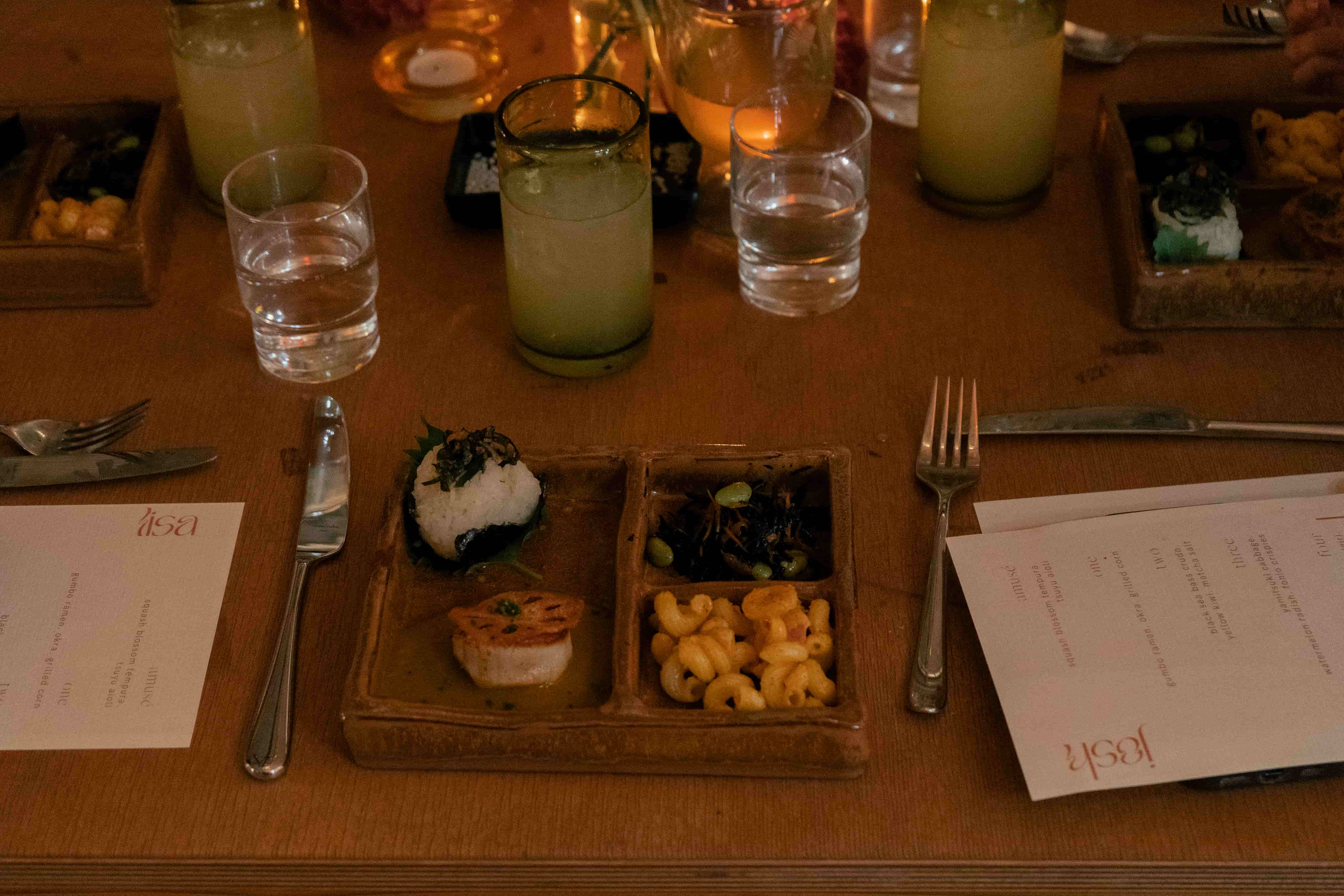A moody table with a serving of food on a coffered plate, with glasses, silverware, and a menu.