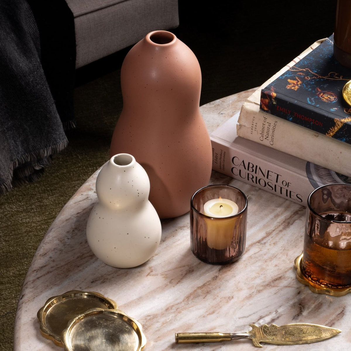 Two gourd-shaped ceramic vases in cream and terracotta sit on a marble coffee table beside a candle, amber glass, brass coasters, and stacked art books.