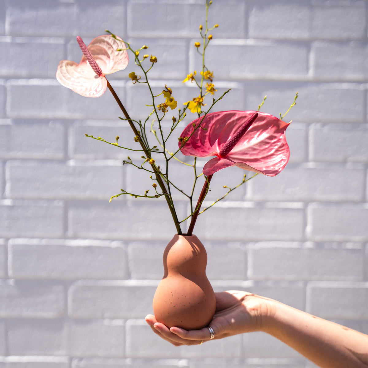 A hand holds out a peach-colored gourd-shaped ceramic vase holding a minimal floral arrangement of two pink anthuriums with bright yellow sprigs, set against a white brick wall.