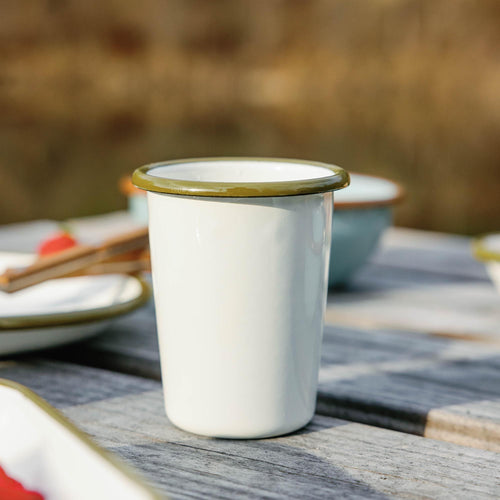 Small white enamel tumbler with an olive green rim sitting on a weathered wooden deck outdoors.