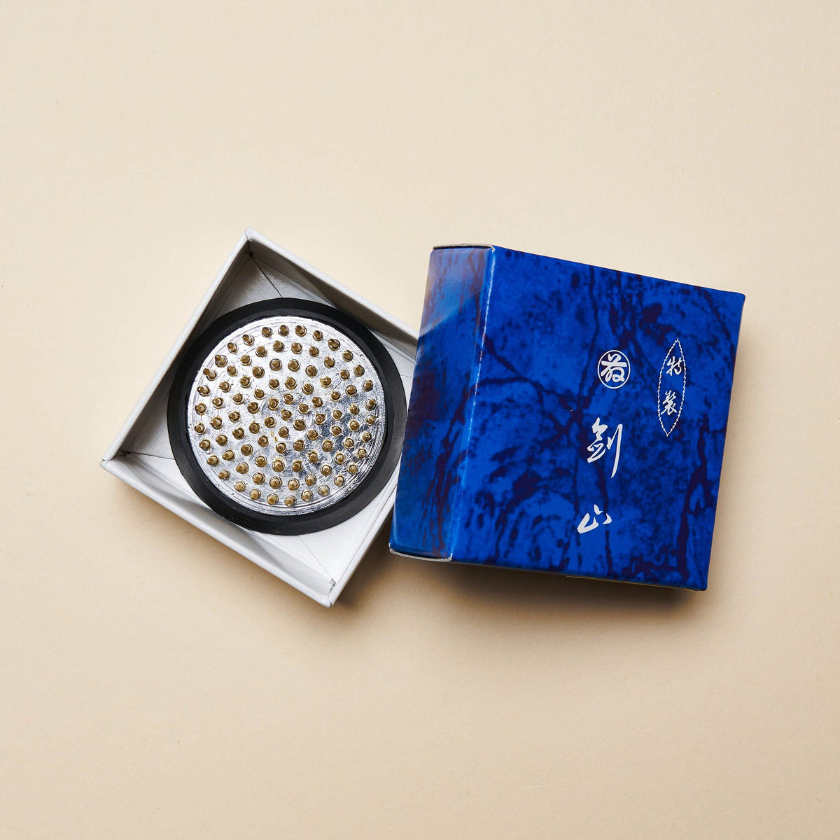 A small "kenzan" – a pin frog made of brass spikes on a flat circular base to assist in flower arrangements sitting inside a white box with a blue top on the right