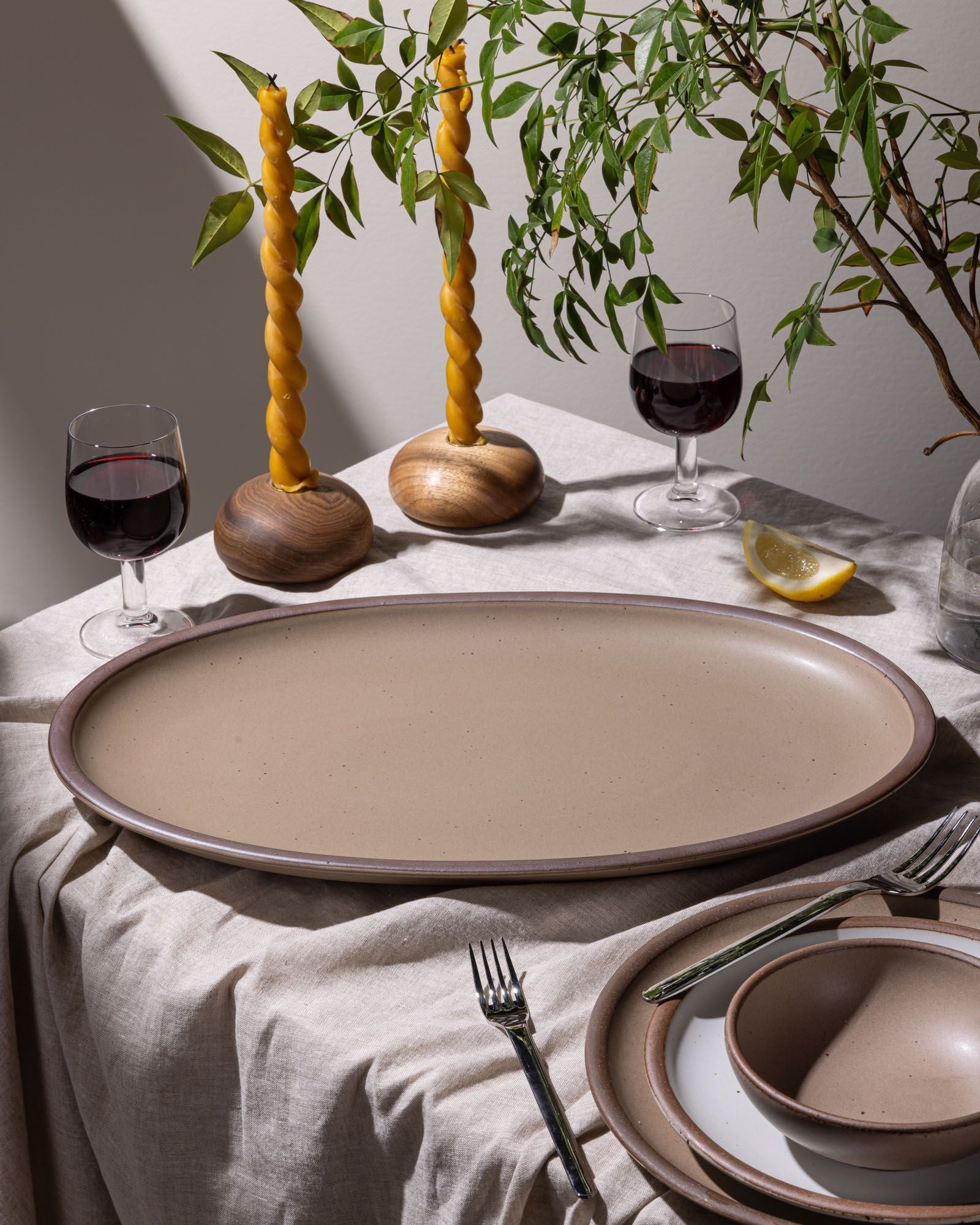 An oval platter in a warm tan brown sits on a table with wine glasses, taper candles, and a plant