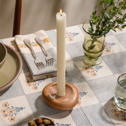 Lit white taper candle in a wooden holder on a patterned table setting with glassware and olives.