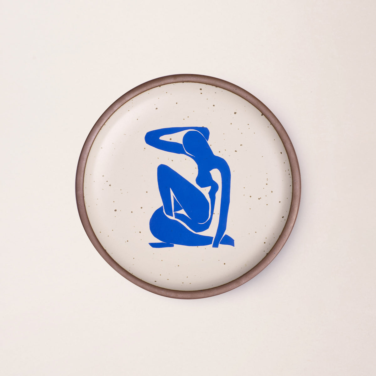 A plate featuring Nu Bleu I, by Henri Matisse, a bright blue, abstract, silhouetted female figure in a curled, seated position, with sharp cut-out edges, centered on an off-white ceramic plate with an unglazed rim.