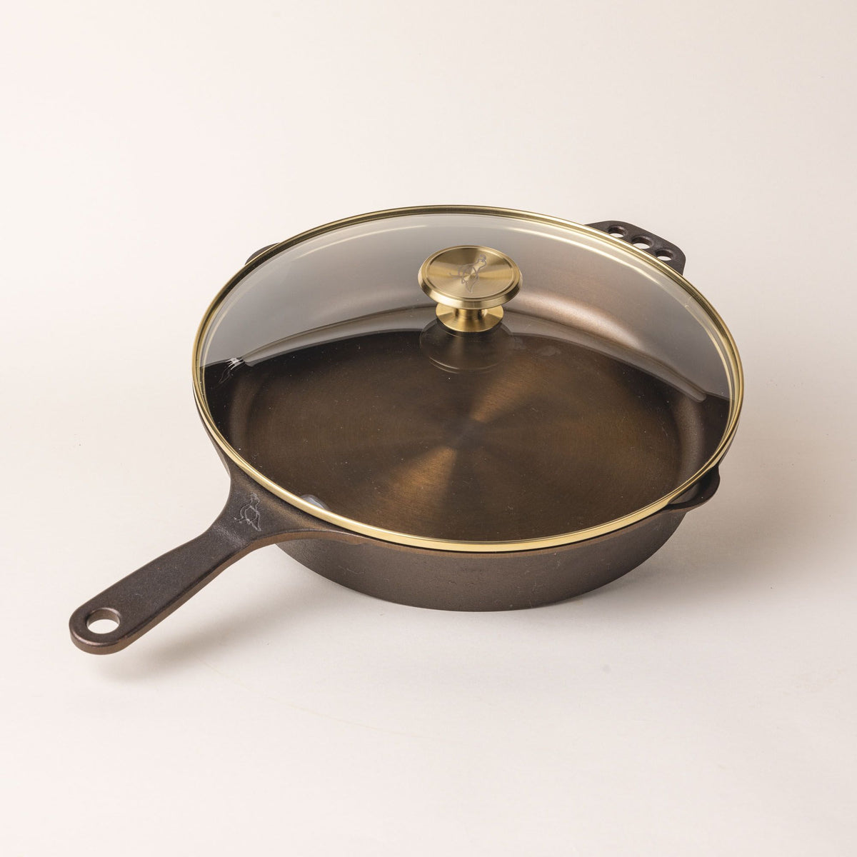 A glass skillet lid with a brass lip and brass handle knob over a cast iron skillet