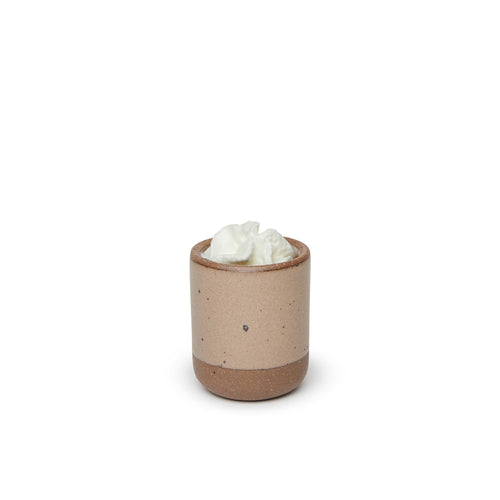 A small, short ceramic mug cup in a warm pale brown color featuring iron speckles and unglazed rim and bottom base, topped with whipped cream.