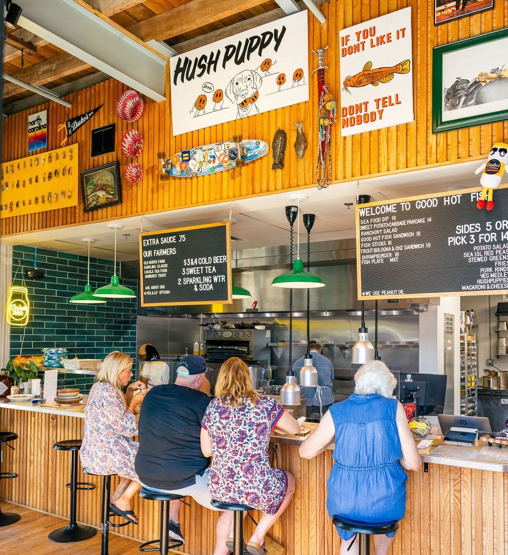 A counter at a restaurant kitchen is filled with people with colorful artwork and a menu above them