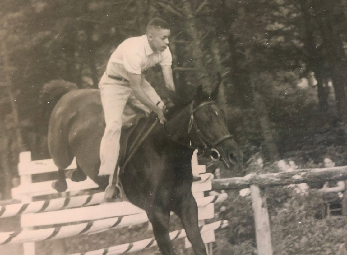 Black and white image of a man riding a horse over a fence 