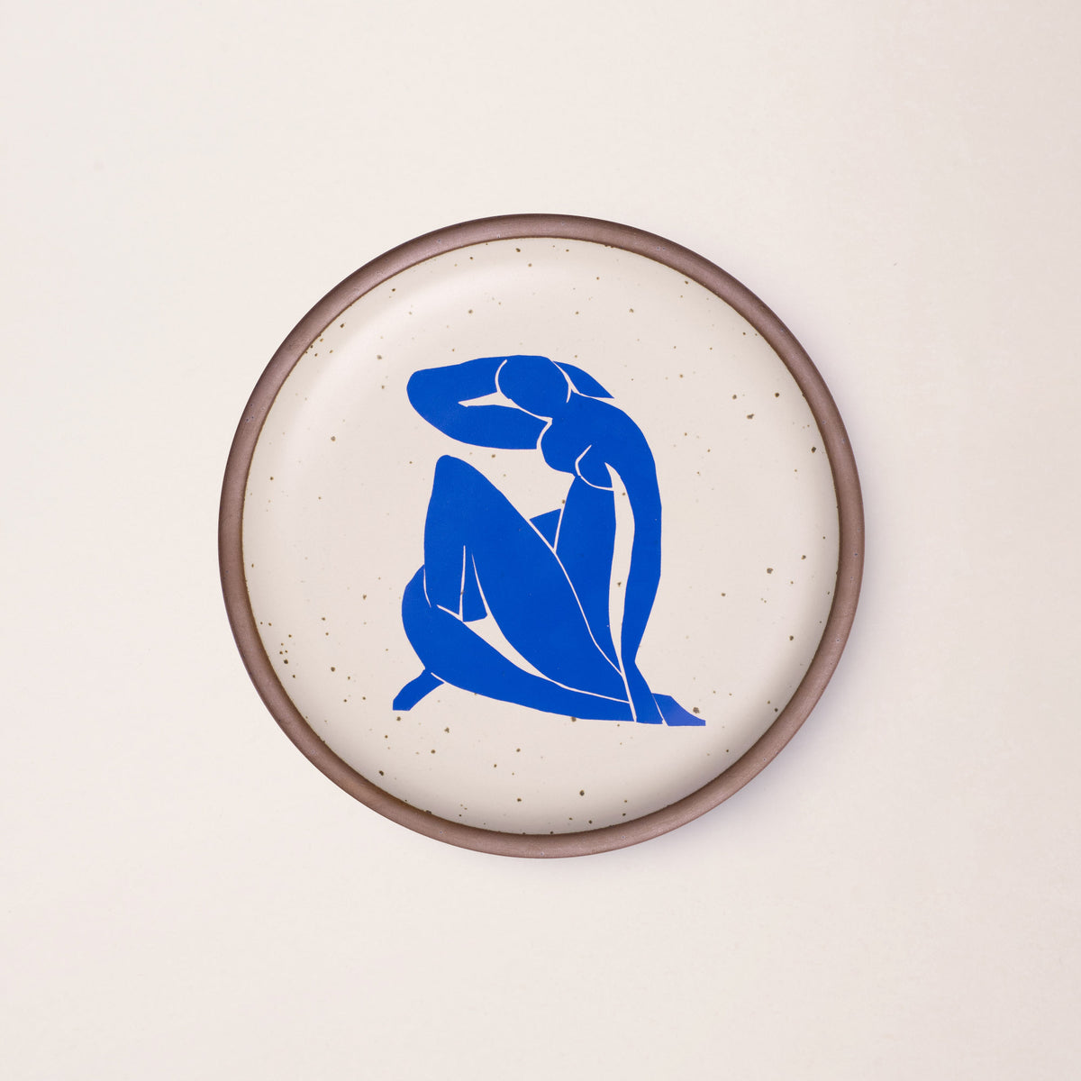 A plate featuring Nu Bleu II, by Henri Matisse, a bright blue, abstract, silhouetted female figure in a curled, seated position, with sharp cut-out edges, centered on an off-white ceramic plate with an unglazed rim.