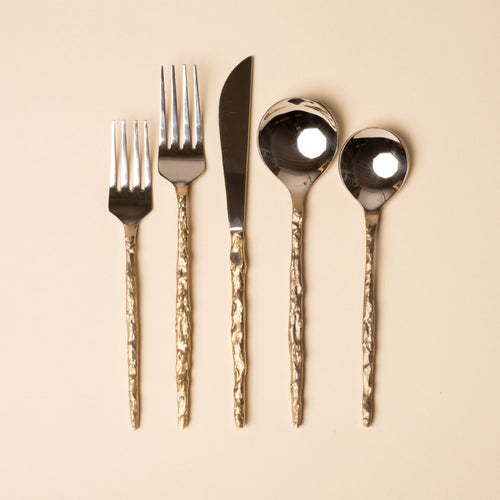 Shiny silver flatware set with hammered gold handles arranged on a beige background.