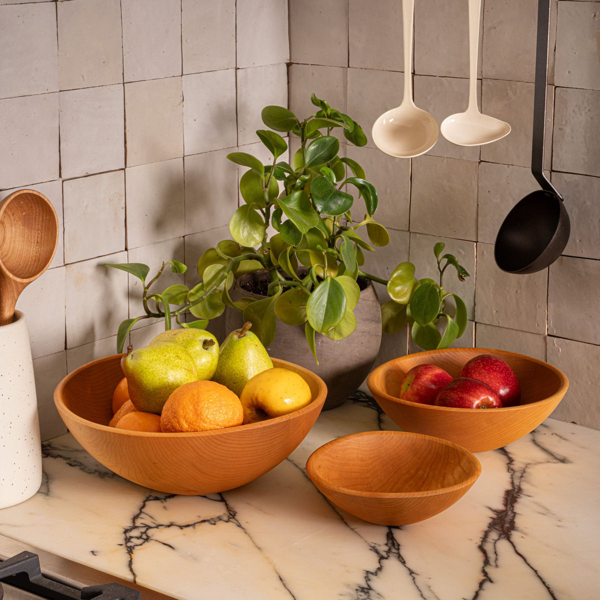 Wooden bowls filled with pears, oranges, and apples on a marble countertop beside a leafy potted plant and hanging kitchen utensils.