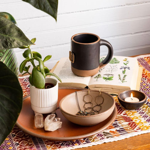 A side table featuring a black ceramic mug sitting on an open book, a tiny bowl holding a tea light, and a shallow bowl holding jewelry, all surrounded by plants.