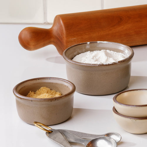 A small and medium ceramic ramekins with flour and sugar, on a countertop with baking accessories.