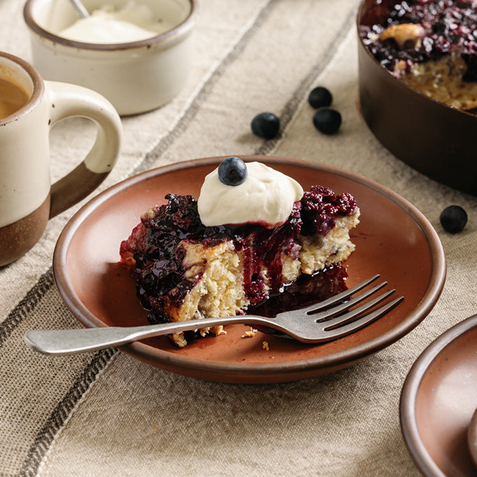 A slice of Blueberry Cake with Blueberry Sauce on a terracotta colored little coupe plate with fork, on a table.