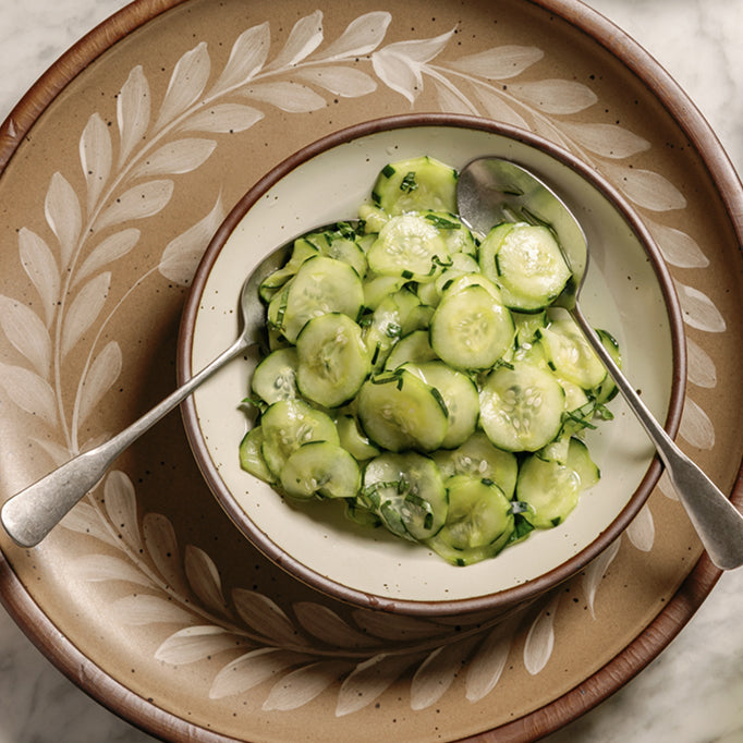A small coupe dish filled with thin sliced cucumbers with spoons, sitting on a hand painted plate.