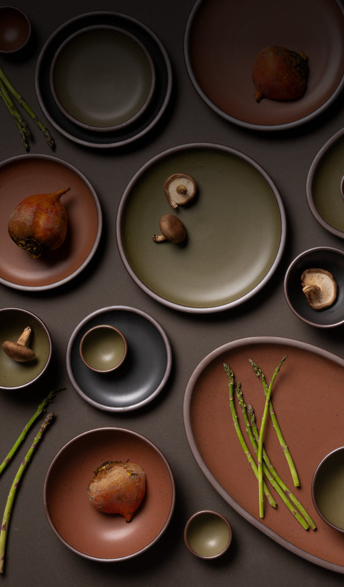 An overhead view of ceramic plates and platters in black, olive, and terracotta colors with food.